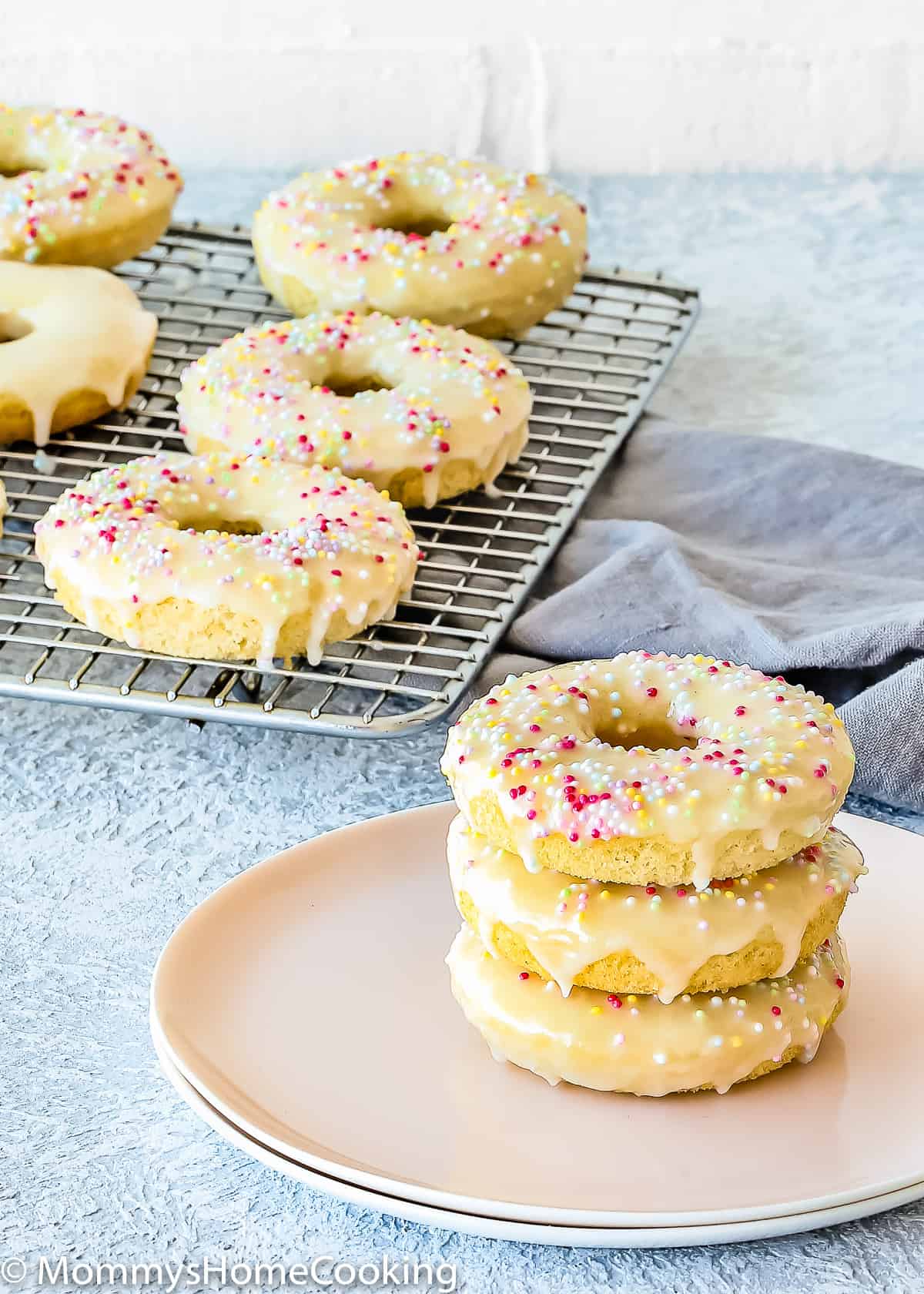 A stack of three donuts on a plate in front of a cooling rack with six more.