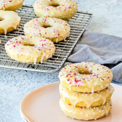 A stack of three donuts on a plate in front of a cooling rack with six more.