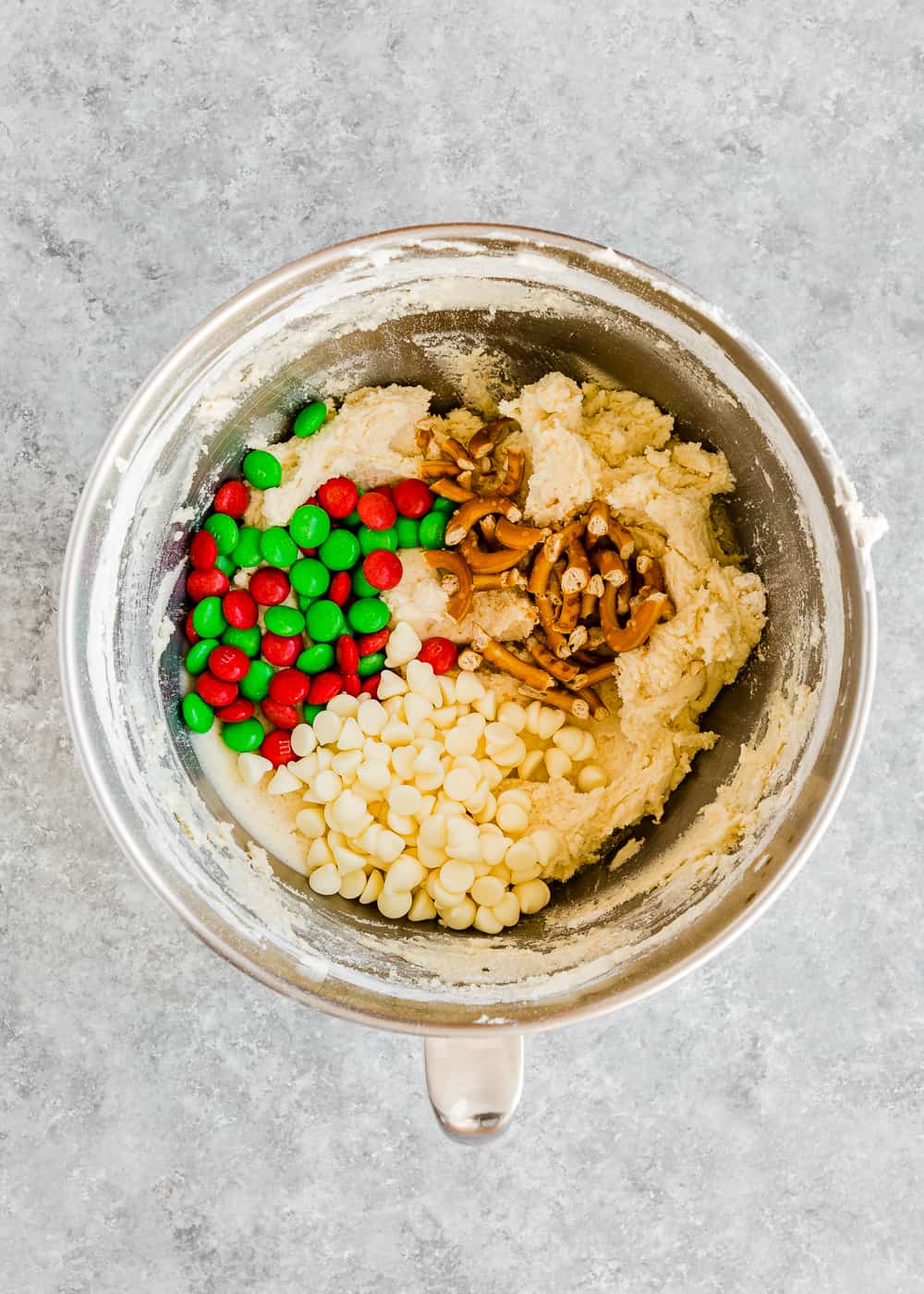 egg-free cookie dough in a stand mixer bowl with m&ms, white chocolate chips and pretzels on top. 