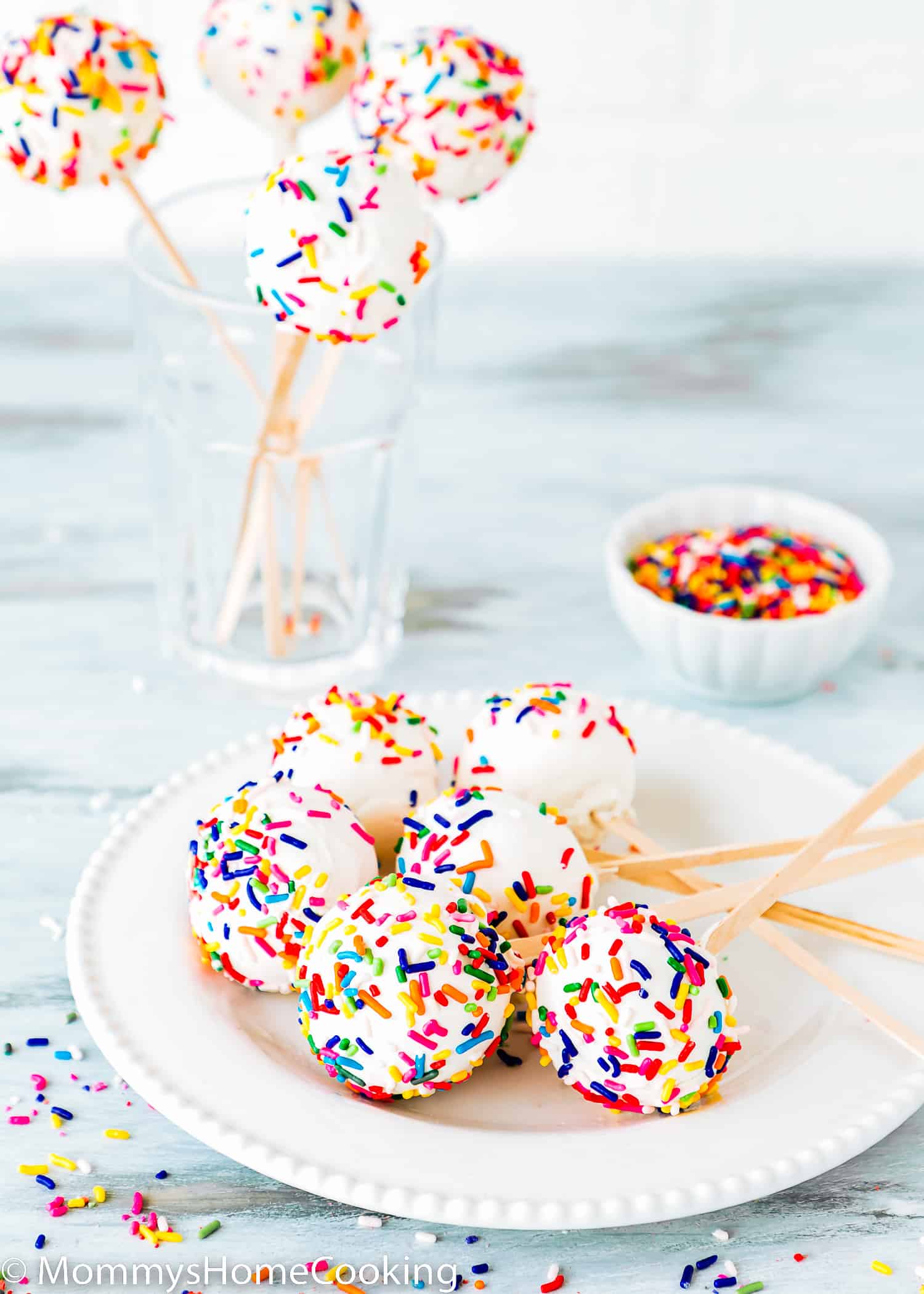 homemade eggless cake pops covered with sprinkles on a white plate with a small bowl of sprinkles and more cake pops in the background.