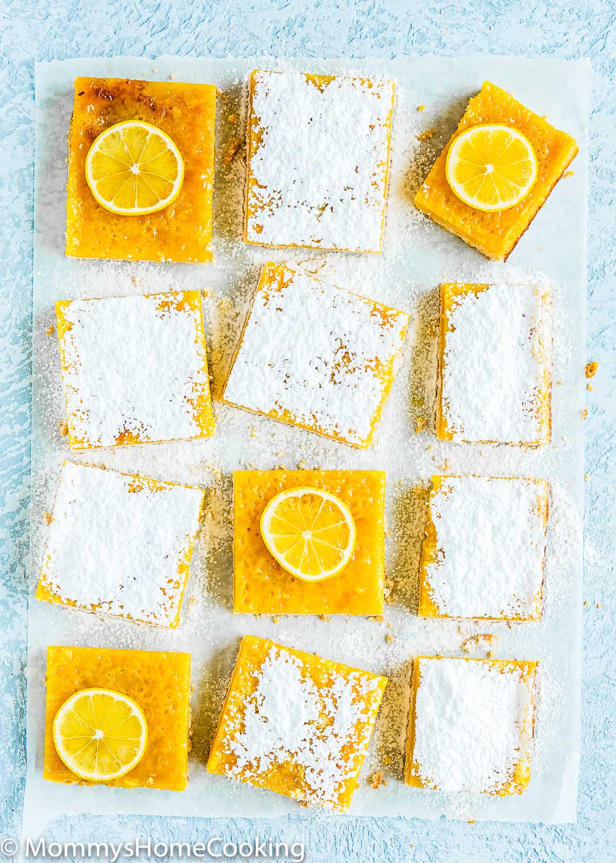 Eggless Lemon Bars with powdered sugar and lemon slices on top over a blue surface