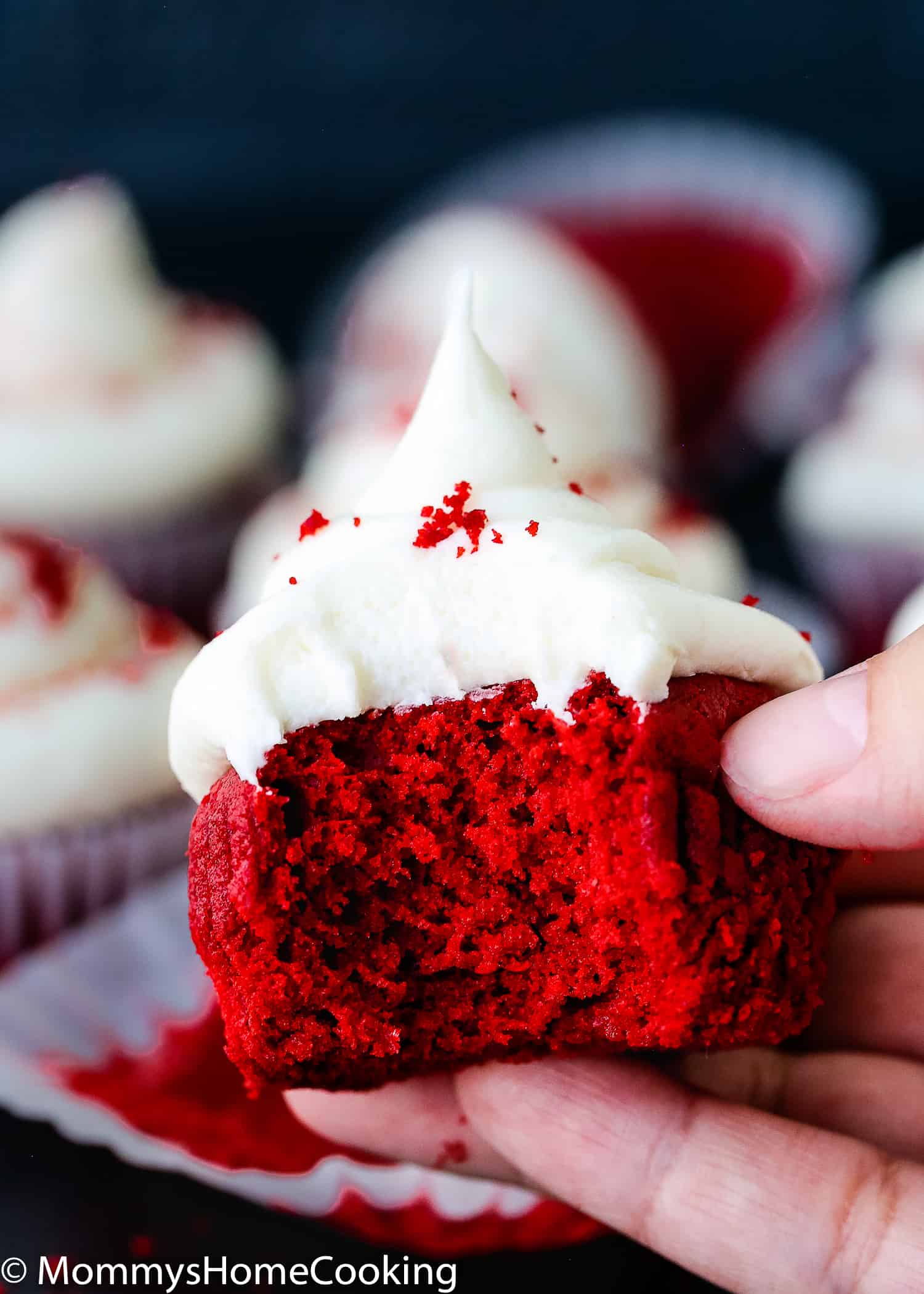 A hand holding a bitten eggless red velvet cupcake showing fluffy inside texture.