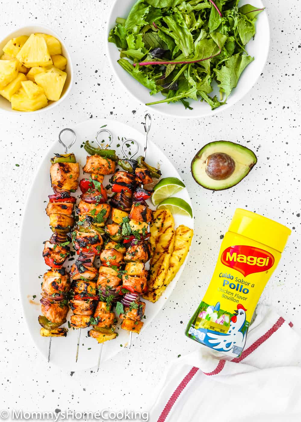 Easy Grilled Hawaiian Chicken Skewers in a white plate with chopped fresh pineapple, green salad, avocado and a granulated chicken Bouillon bottle