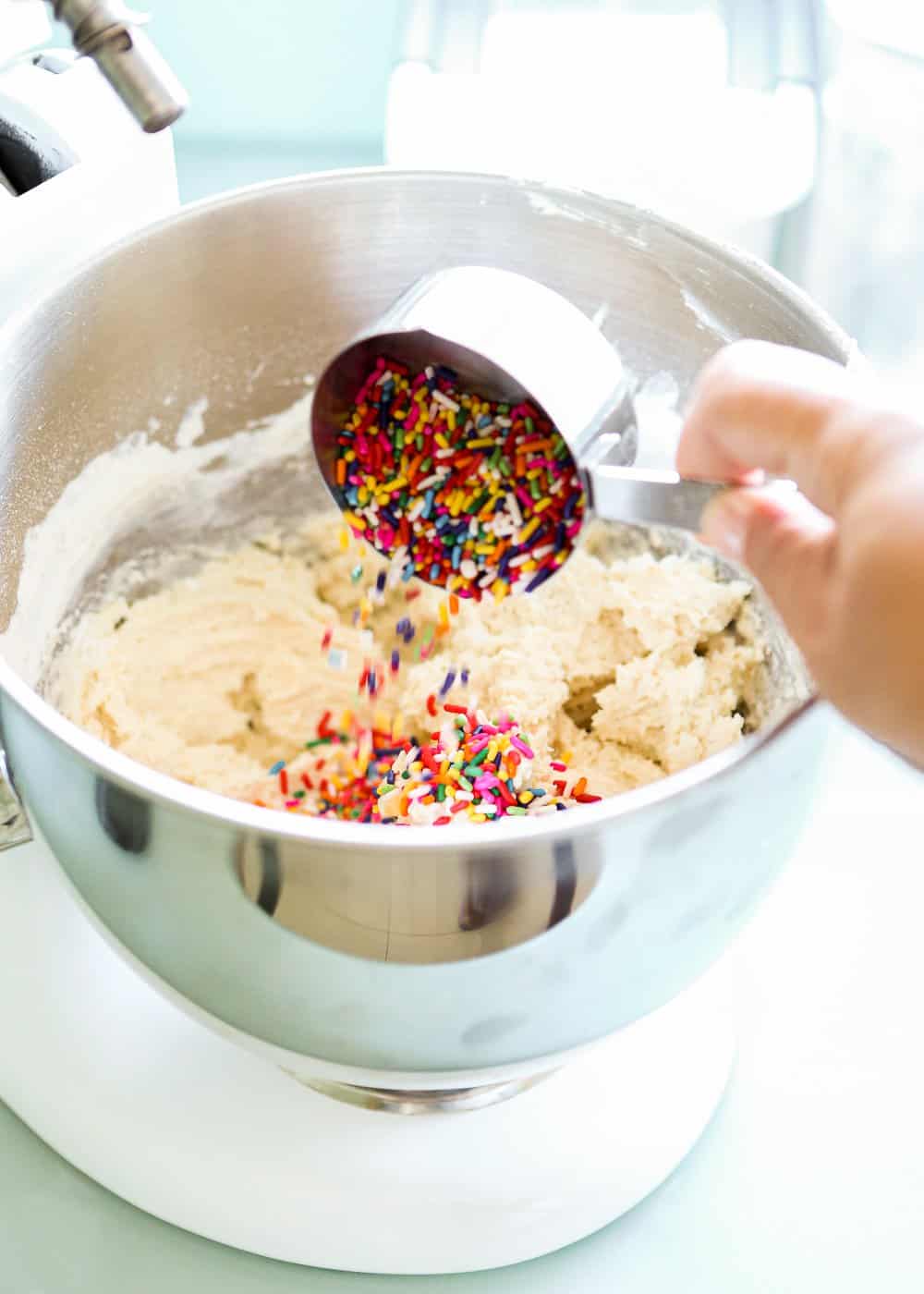 a hand adding sprinkles to an egg-free cookie dough in a bowl of a stand mixer. 