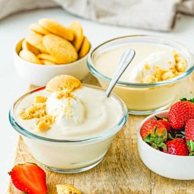 two bowls with Egg free homemade vanilla pudding with whipped cream over a wooden board with fresh strawberries, more vanilla wafers and a gray kitchen towel.