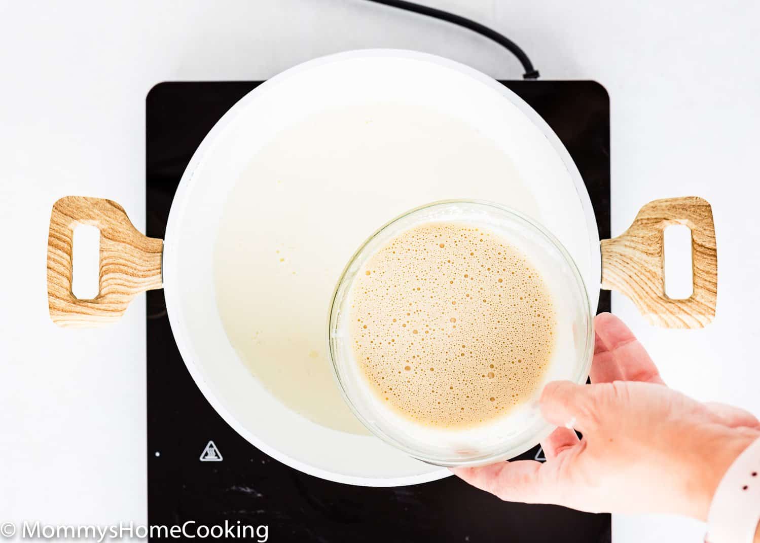 a hand with a bowl of evaporated milk over a pot with milk.