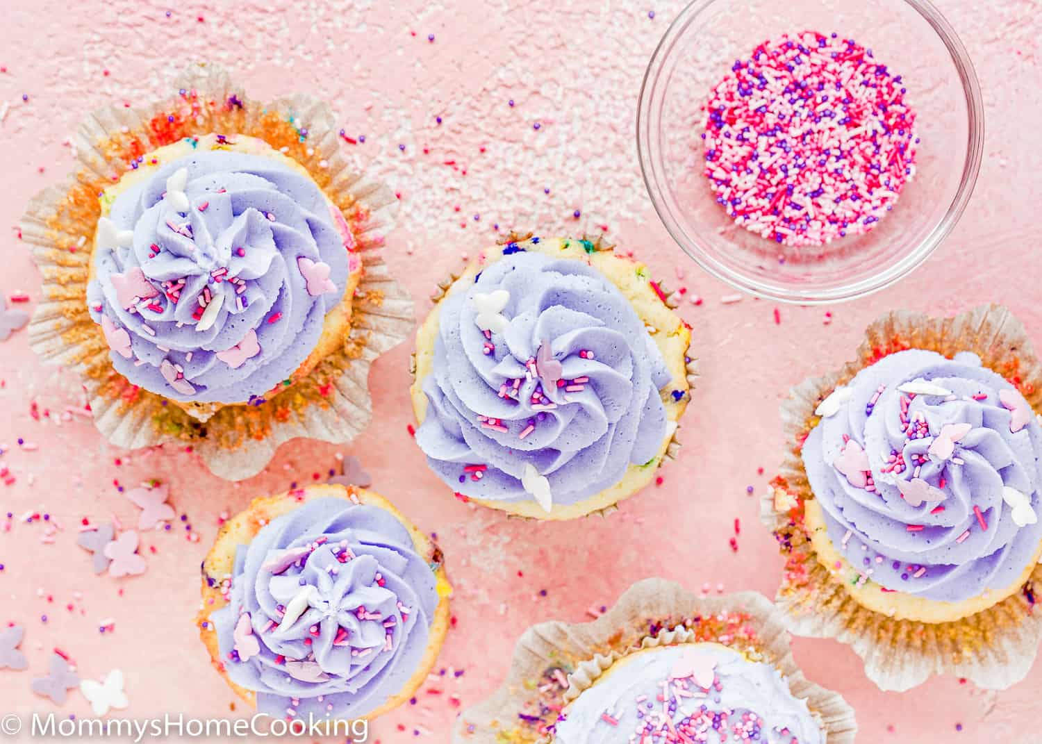Eggless Funfetti Cupcakes over a pink surface with sprinkles and butter cream on top.