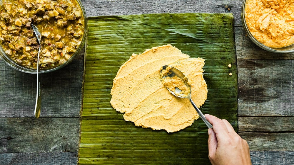 a hand holding a spoon and spreading hallaca dough onto a banana leave. 