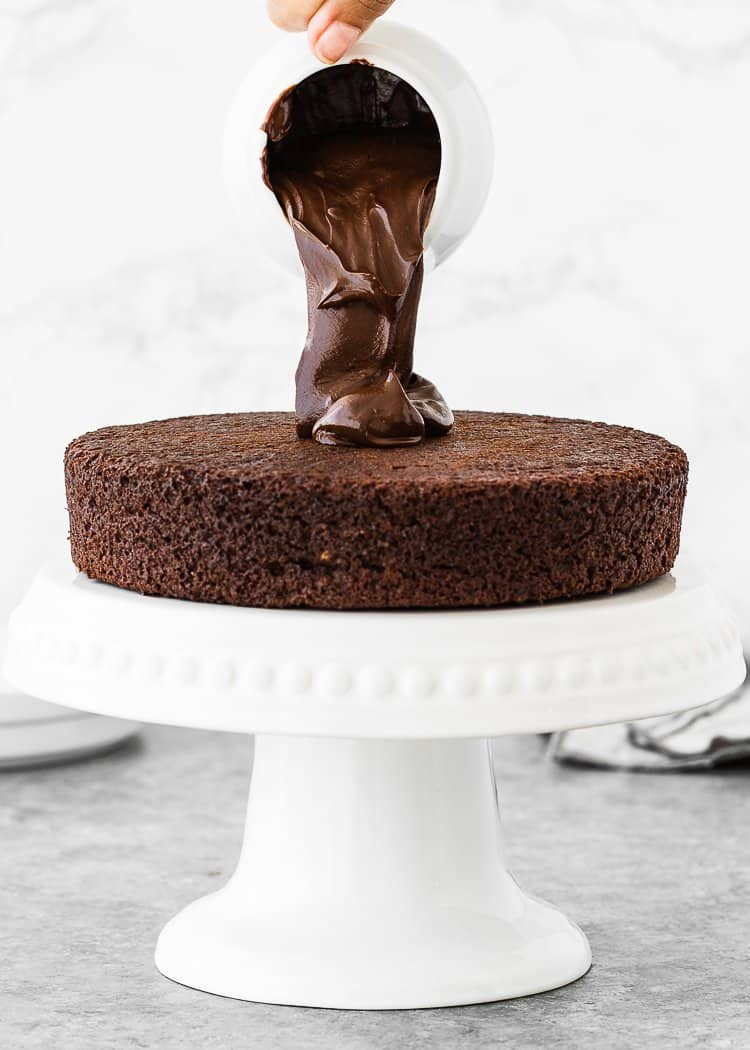 chocolate ganache being poured over an egg-free chocolate cake layer over a cake stand. 