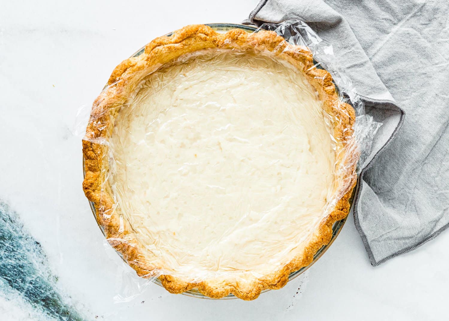 Eggless Coconut Cream Pie in a pie dish covered with plastic wrap with a grey kitchen towel on the side. 