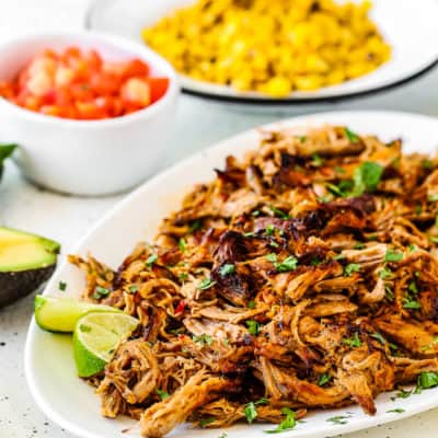 Slow Cooker Chipotle Carnitas in a serving plate with tomatoes and corn on the background.