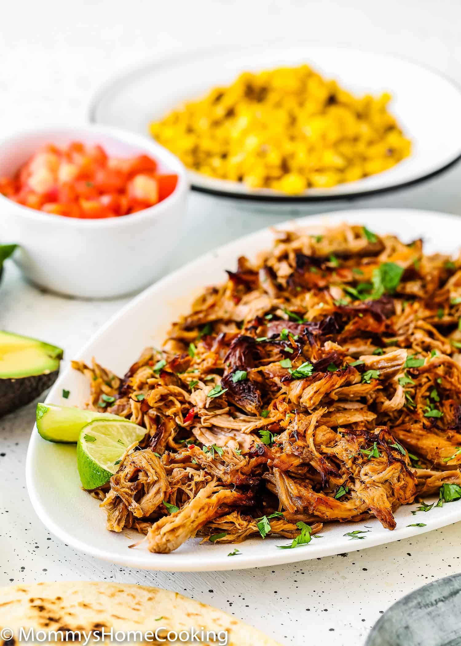 Slow Cooker Chipotle Carnitas in a serving plate with tomatoes and corn on the background.