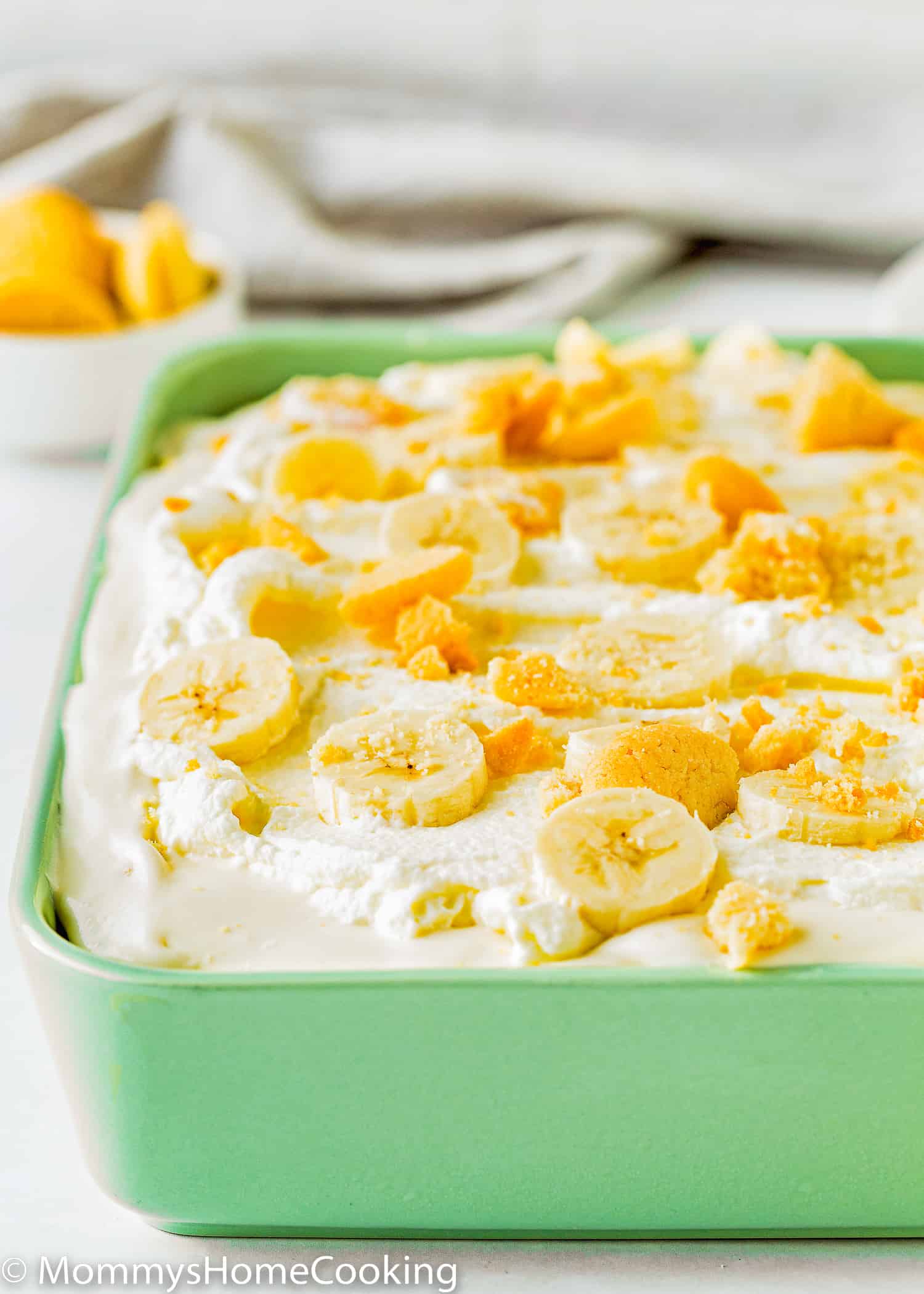 Eggless banana pudding in a green rectangular dish with a kitchen towel and a small bowl with wafers on the background.