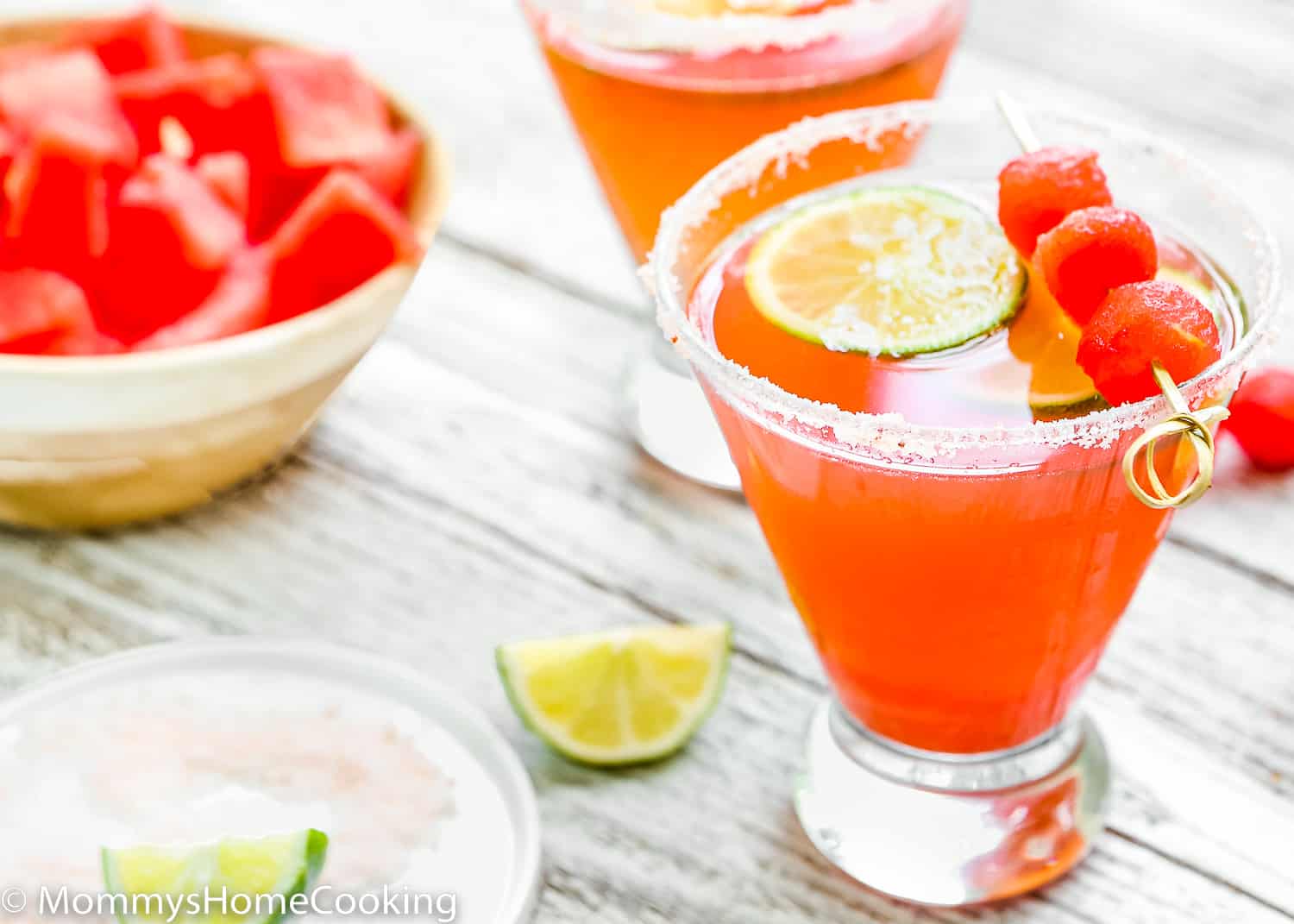 two Watermelon Cocktails over a wooden surface surrounded by lemon slices and a bowl of cubed watermelon.